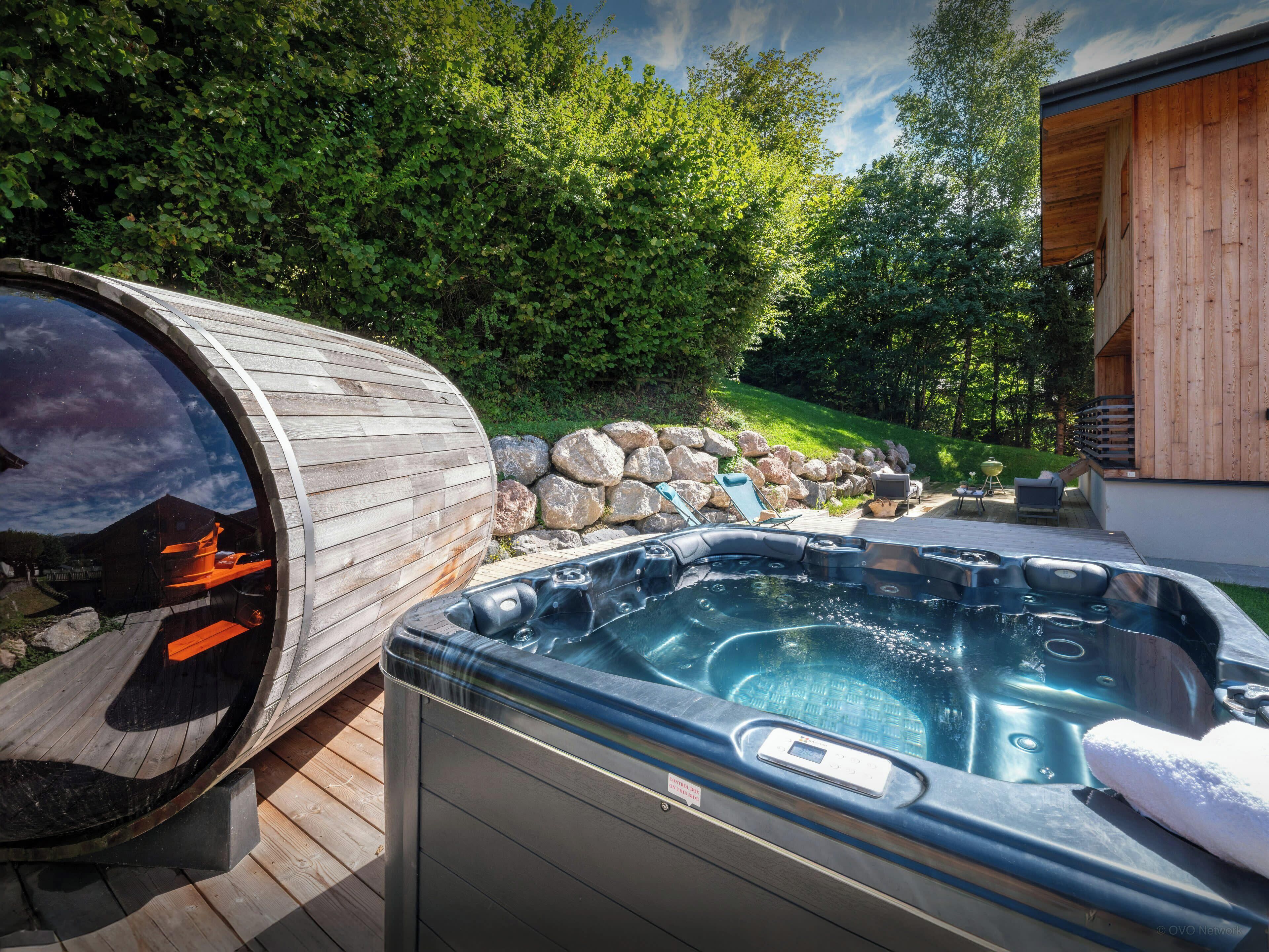Un bain à remous et un sauna sur une terrasse en bois, avec un mur de pierres et de la verdure en arrière-plan.