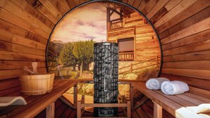 A sauna with a wooden interior, a stone heater, wooden benches, and a view of a wooden cabin and trees.
