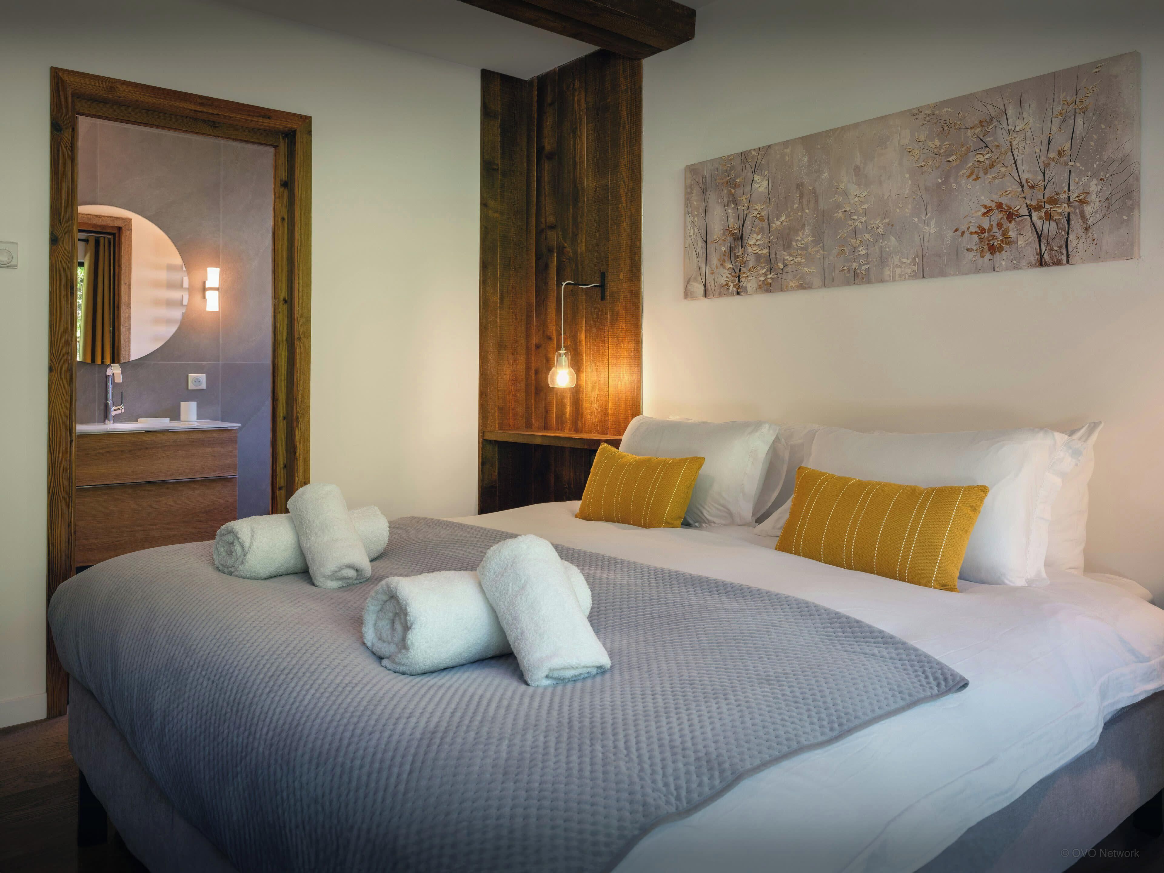 Een keurig opgemaakt bed met witte en gele kussens, een grijze deken en opgerolde handdoeken. Er is een houten hoofdbord, een aan de muur hangend schilderij van bomen, en een houten vloer.