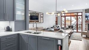 A modern kitchen with grey cabinets, a large island, and a stone fireplace. - Luxurious Home In The Village Core - Hot Tub! (Sun Peaks)