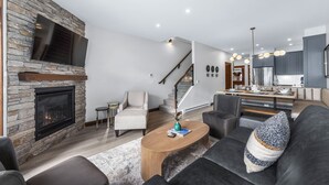 A modern living room with a stone fireplace, a sofa, a coffee table, and a kitchen area in the background. - Luxurious Home In The Village Core - Hot Tub! (Sun Peaks)