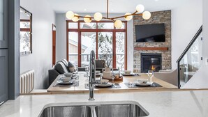 A modern kitchen with a stone fireplace, large windows, and a dining area set up for a meal. - Luxurious Home In The Village Core - Hot Tub! (Sun Peaks)