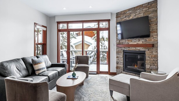 A modern living room with a stone fireplace, a flat-screen TV, and large windows with a view of snow-covered trees. - Luxurious Home In The Village Core - Hot Tub! (Sun Peaks)