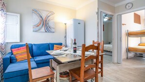 A compact living space with a blue sofa, wooden dining table, and a bunk bed in the background.
