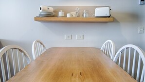 Ein Esstisch mit vier weißen Stühlen, ein Holzregal darüber mit Tellern, Tassen und einem Toaster, sowie zwei Steckdosen an der Wand.