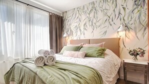 Una cama bien tendida con toallas enrolladas, una manta verde y varios almohadones. Se observa una lámpara de pared y un florero con flores.