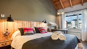 A bedroom with a wooden headboard, a bed with multiple pillows, a bedside table with a lamp, a window with a view of buildings, and a radiator.