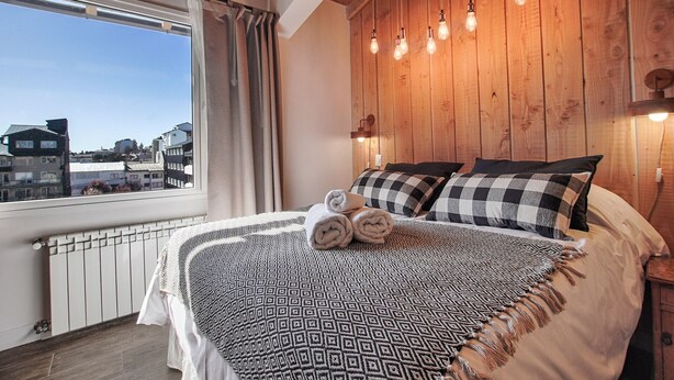 A bedroom with a large bed, checkered pillows, a view of the city through a window, and wooden paneling on the walls.