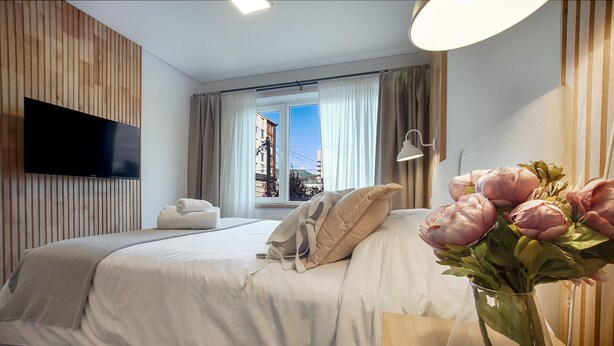 A modern hotel room with a bed, a TV, a window with curtains, and a vase with flowers.
