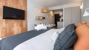 Une chambre d’hôtel moderne avec un grand lit, un téléviseur à écran plat fixé au mur, un coin repas avec une table dressée pour deux personnes, et une kitchenette à l’arrière-plan.