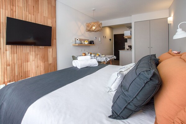 Una habitación de hotel moderna con una cama grande, un televisor de pantalla plana en la pared, un rincón de comedor con una mesa para dos personas y una kitchenette al fondo.