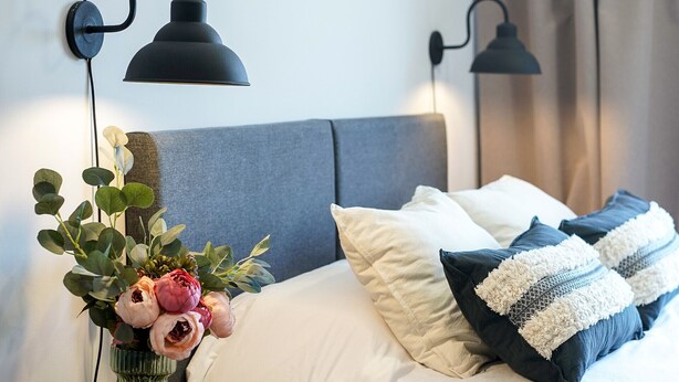 Una cama bien hecha con almohadas blancas y azules, un jarrón con flores y dos lámparas de pared.
