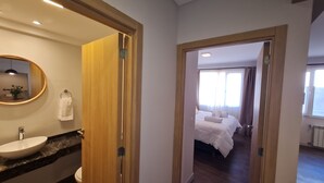 A hotel room with a wooden door leading to a bathroom with a sink and mirror, a bed with white bedding, and a window with curtains.