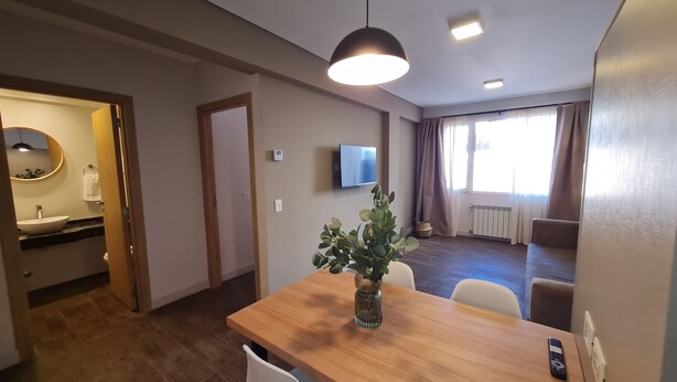 A modern hotel room with a wooden dining table, a vase of green plants, a flat-screen TV mounted on the wall, and a large window with curtains.