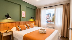Hotellihuone, jossa on sänky, yöpöytä, lamppu, ikkuna verhoilla ja näkymä ulkona oleviin rakennuksiin.