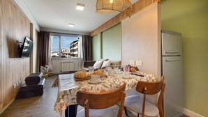 Ein Essbereich mit Tisch für zwei Personen, einer Couch, einem Fernseher und Blick auf Gebäude.