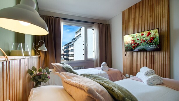 Ein Hotelzimmer mit zwei Betten, einem an der Wand befestigten Fernseher und einem Fenster mit Blick auf Gebäude.