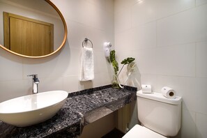 Une salle de bain avec un lavabo blanc, un miroir rond, une serviette, des toilettes et une plante.