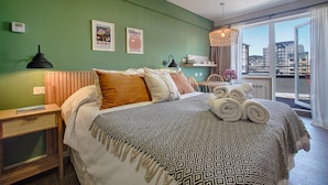 Een moderne hotelkamer met een groene wand, een bed met behang, een nachtkastje, een lamp en uitzicht op de stad door grote ramen.