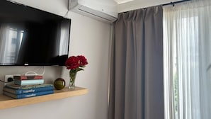Habitación con televisión de pantalla plana en la pared, un equipo de aire acondicionado encima, una ventana con cortinas translúcidas y un estante con libros y un florero.