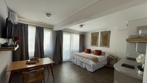 Habitación de hotel con cama, escritorio, silla, mesita, zona de cocina y cortinas en las ventanas.