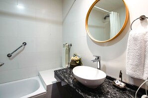 Un baño con lavamanos blanco, un espejo redondo, toallero, cortina de ducha y barra de apoyo.