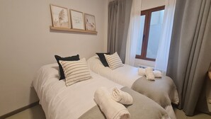 Ein Zimmer mit zwei Betten, jeweils mit Kissen und einem zusammengerollten Handtuch. An den Wänden über den Betten hängen gerahmte Bilder.