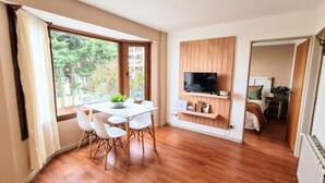 Una habitación moderna con suelo de madera, una mesa blanca con sillas, un televisor montado en un panel de madera y vistas al exterior con vegetación.