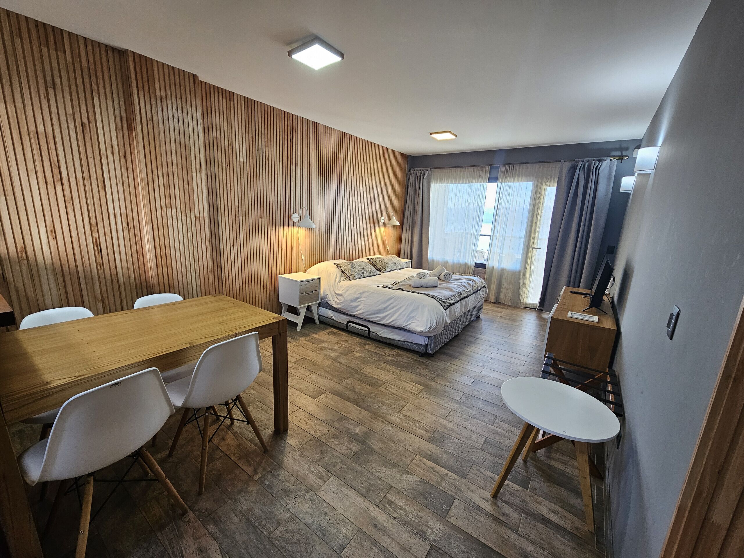 A modern bedroom with a wooden slat wall, a wooden dining table with chairs, a bed with pillows, and a desk with a lamp.