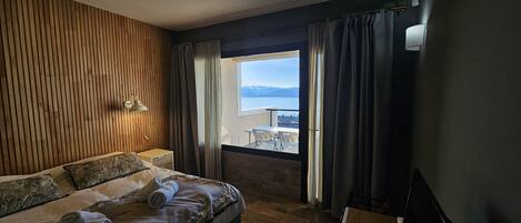 A modern hotel room with a large bed, a desk, a lamp, and a view of the sea.