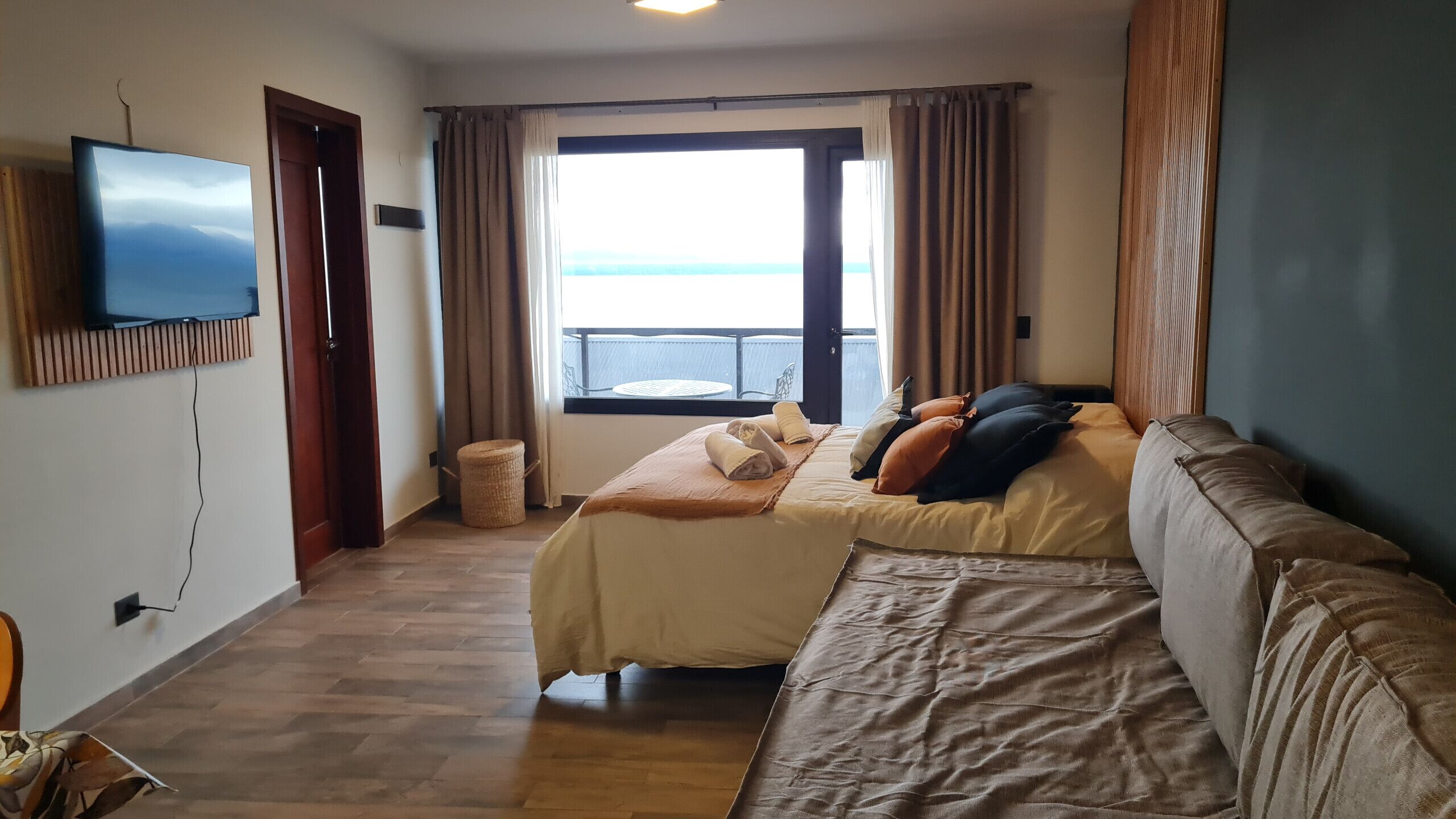 Une chambre d’hôtel avec un lit, un canapé, un téléviseur fixé au mur, une fenêtre avec des rideaux et une petite table ronde.