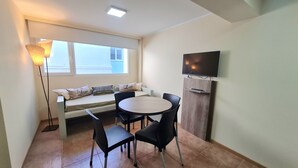Una habitació petita amb una taula rodona, quatre cadires, una banca amb coixins, una làmpada de peu i un televisor fixat a la paret.
