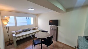 Una habitació petita i acollidora amb una banca al costat de la finestra, una taula rodona amb dues cadires, una televisió plana penjada en un moble de fusta i una làmpada de peu.