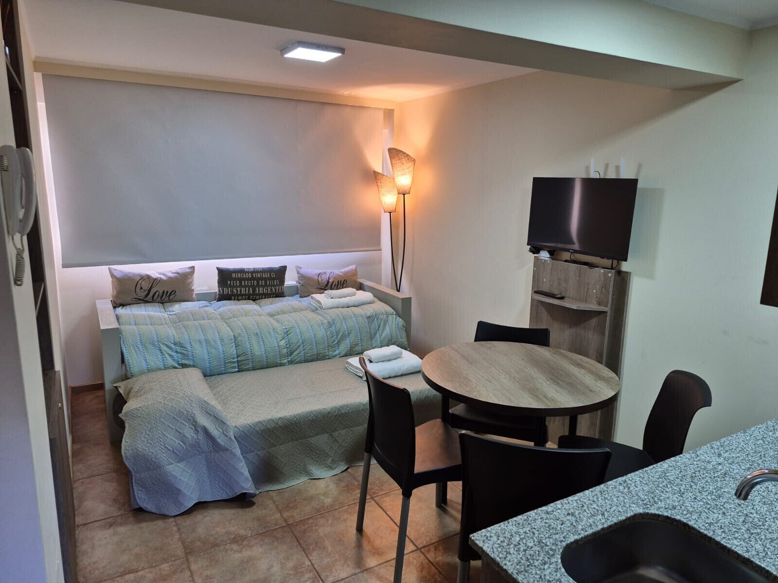 A compact hotel room with a bed, bedside table, chair, and a small dining table with chairs.