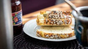 A plate with slices of bread topped with seeds, a jar of jam, and a cup of coffee on a woven mat.