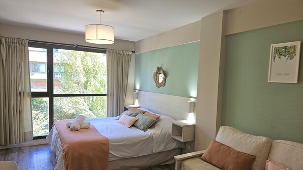 Ein Schlafzimmer mit einem Bett, Nachttischen, einem Sessel, einer Couch, einem Fenster mit Vorhängen und einem Wandbild.