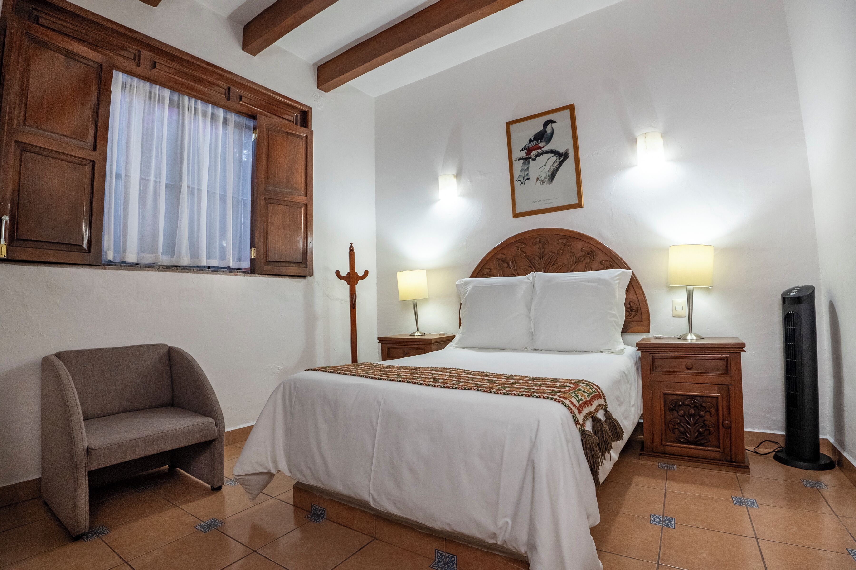 Un dormitorio con una cama, dos lámparas de noche, una mesita de madera, una silla y una ventana con cortinas.