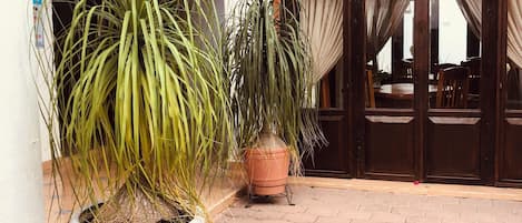 Un patio con plantas en macetas, piso de baldosas y un área de comedor con cortinas.