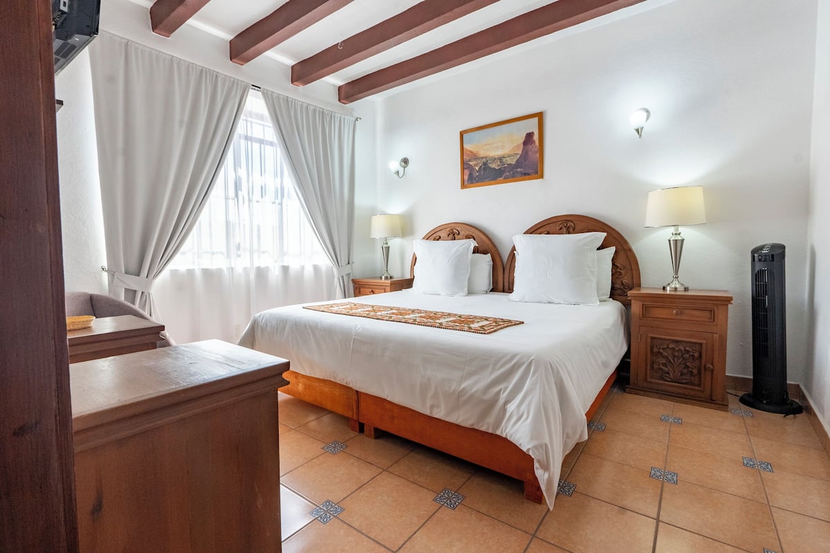 A bedroom with a large bed, wooden headboard, two bedside tables with lamps, a nightstand, a standing heater, and a window with curtains.