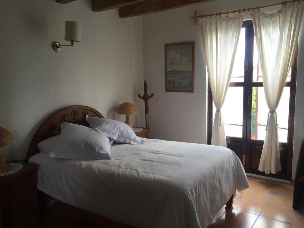 A bedroom with a wooden bed, white bedding, a nightstand with a lamp, a wall-mounted lamp, a window with curtains, and a painting on the wall.