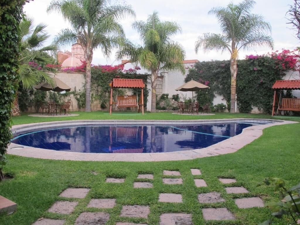 Una piscina rodeada de un césped cuidado, con palmeras y un pabellón al fondo.