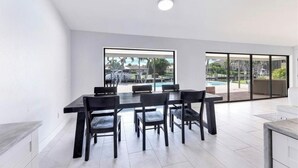 A modern dining area with a large table and chairs, large windows with a view of a swimming pool and greenery.