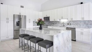 A modern kitchen with white cabinets, a marble countertop, a stainless steel refrigerator, a microwave, a sink, and three bar stools.