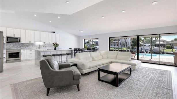 A modern living room with a sectional sofa, a coffee table, and a grey armchair. There is a kitchen area with white cabinets and a microwave. The room has large windows offering a view of a swimming pool and a residential area.