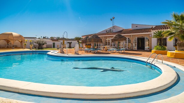 Une piscine ornée d’une statue de dauphin, avec des chaises longues, et un bâtiment à l’arrière-plan, dont le toit est recouvert de tuiles.