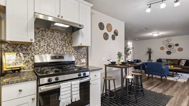 A modern kitchen with white cabinets, a stainless steel stove, and a tiled backsplash. There is a dining area with stools and a blue sofa in the background. - Incredible Stay Fast Wifi Fully Equipped (San Antonio)