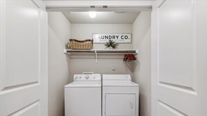 A laundry room with two white washers and dryers, a sign above them reading "LAUNDRY CO. WASH & FOLD", and a shelf with baskets and a plant. - Incredible Stay Fast Wifi Fully Equipped (San Antonio)
