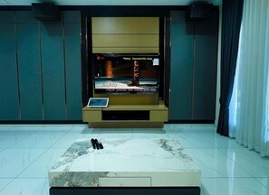 A modern hotel room with a large flat-screen TV mounted on a wall, a small table with a laptop, and a marble-patterned area rug. - Villa Chee (Sekinchan)