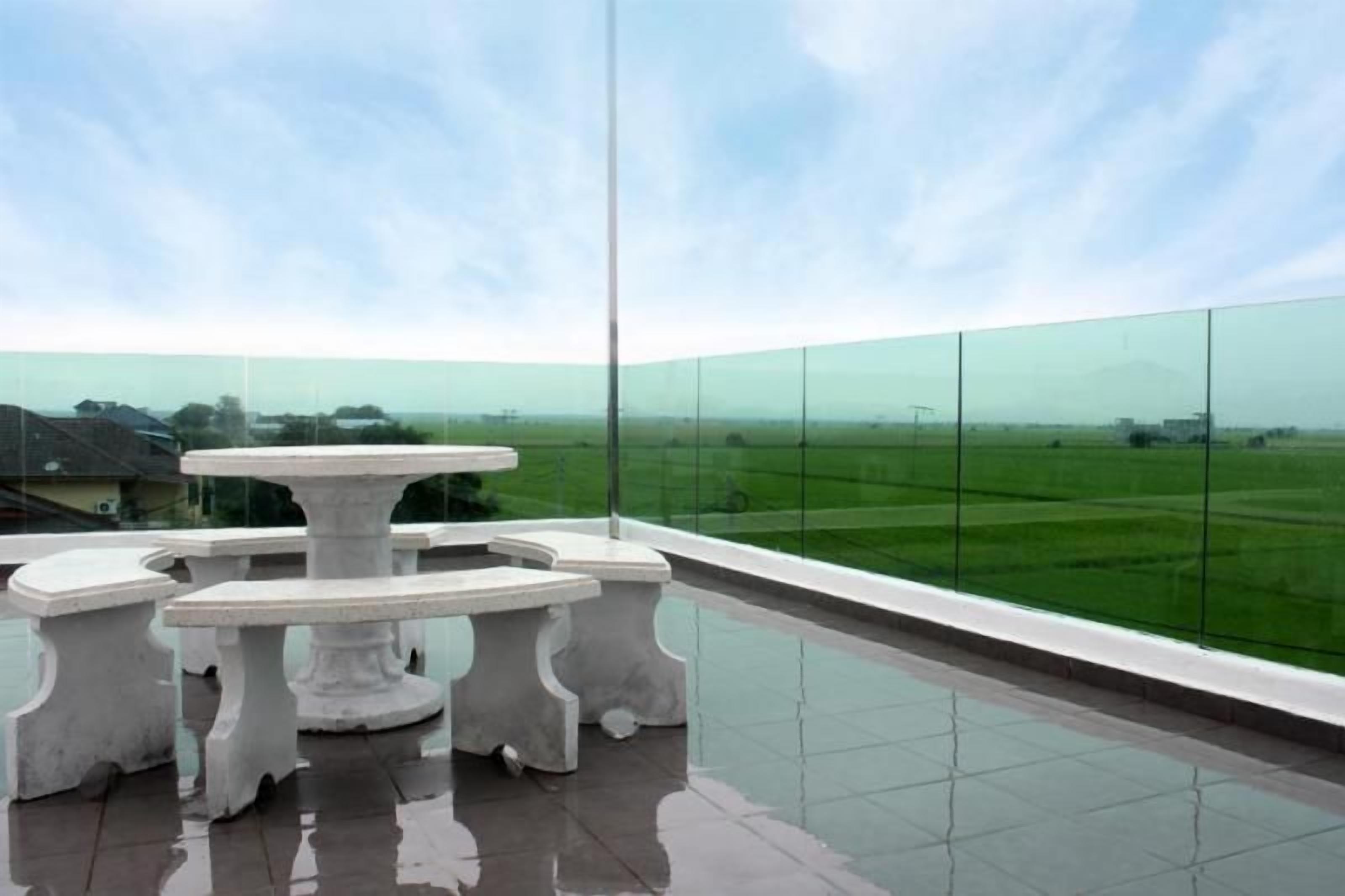 Un balcón con barandilla de vidrio, una mesa blanca y vistas a un paisaje verde.
