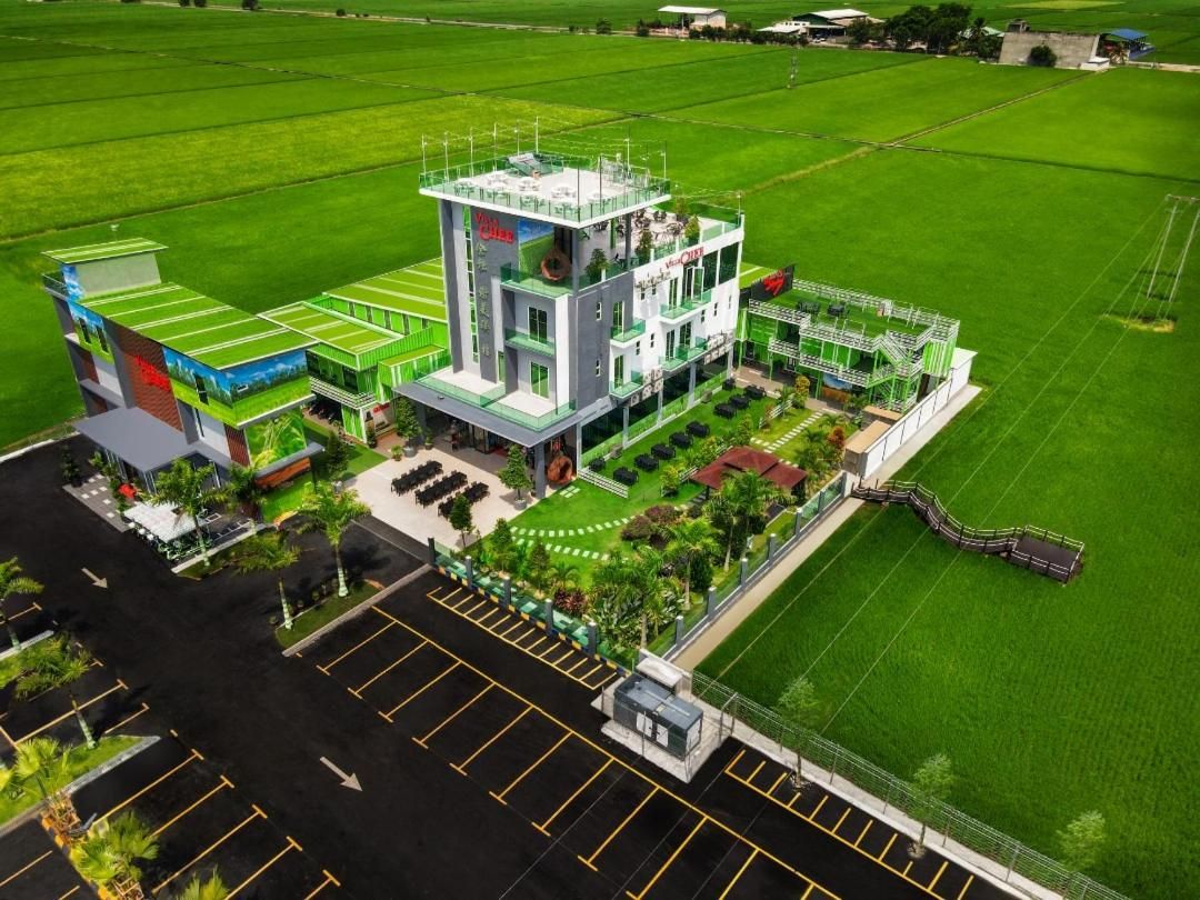 Un edificio moderno con techo plano, rodeado de campos verdes y un estacionamiento.